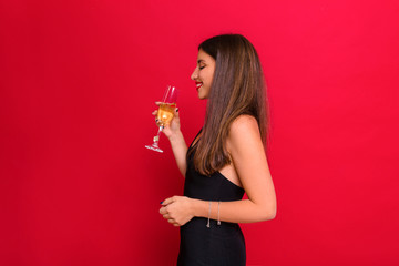 Close up portrait pretty lady on red background celebrating new year, wearing black dress  drinking champagne and celebration holidays, sparkling confetti, smiling to camera.