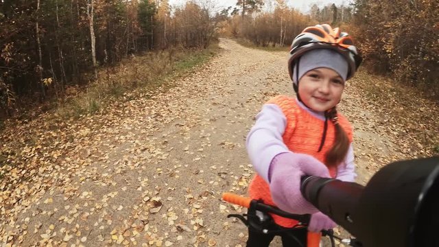 Take selfie on camera phone. One caucasian children rides bike road in autumn park. Girl riding black orange cycle in forest. Kid goes do bicycle sports. Biker motion ride with backpack and helmet.