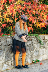 Fototapeta na wymiar Outdoor portrait of adorable little red-haired girl, posing next to bright tree with orange leaves, wearing check jacket, black skirt and hat