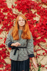 Autumn portrait of adorable red-haired preteen girl girl check jacket and glasses, posing next to wall with red ivy leaves, fall fashion for kids