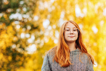 Outdoor close up portrait of beautiful red-haired preteen girl wearing check coat and eyeglasses
