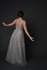Fototapeta premium full length portrait of brunette girl wearing beautiful long gown, standing pose with background to the camera on grey studio background.
