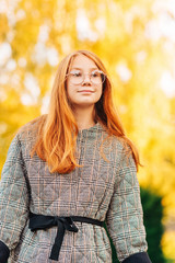 Outdoor close up portrait of beautiful red-haired preteen girl wearing check coat and eyeglasses