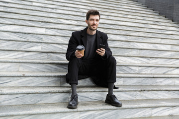 Handsome young business man sitting outdoors using mobile phone drinking coffee.