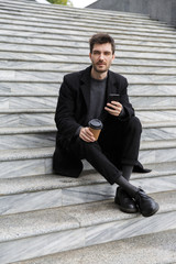 Handsome young business man sitting outdoors using mobile phone drinking coffee.