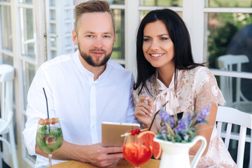 Close-up of happy lovers are reviewing photos from their last trip, sitting in a cafe and enjoying a tablet. Smiling blonde guy and beautiful brunette use gadgets.