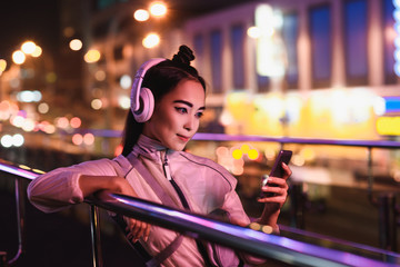 attractive asian girl listening music with smartphone on street with neon light in evening, city of...