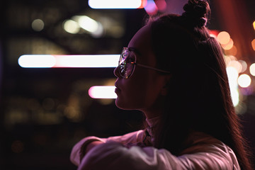 Obraz premium side view of stylish asian girl in glasses looking away on street with neon light in evening, city of future concept