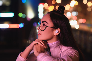 side view of stylish asian girl in glasses and wireless earphone looking away on street with neon light in evening, city of future concept