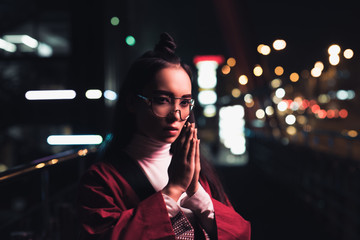beautiful asian girl in burgundy kimono making namaste gesture on street with neon light in...