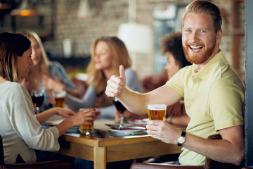 Man drinking beer and giving thumbs up while sitting in restaurant. In background his friends chatting and drinking alcohol.