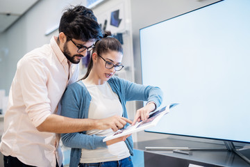 Couple holding catalog and pointing at smart TV they like. New technologies concept.