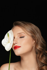 Obraz na płótnie Canvas Portrait of young attractive lady with wavy hair and red lips dreamily covering eye with white calla flower over black background isolated