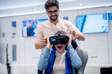 Amazed woman looking through VR glasses while sitting on interactive VR seat. Man standing behind her back.
