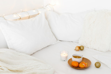 White cozy bedroom with holiday decoration. Bed with white bedding set, pillows, wooden plate, cup of tea, tangerines, christmas balls, candle and garland lights. Home christmas decor.