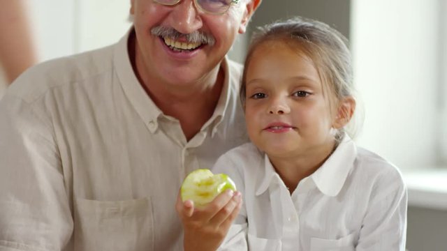 Pretty little girl sitting on knees of grandfather, eating green apple and opening her mouth while watching exciting TV program during morning breakfast at home