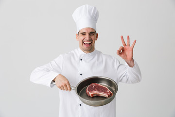 Excited young man chef indoors isolated over white wall background holding frying pan cooking meat.