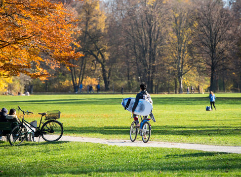 Englischer Garten M&uuml;nchen - Surfer auf Rad f&auml;hrt l&auml;&szlig;ig mit Brett zur Eisbachwelle