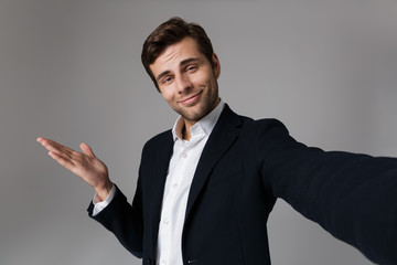 Image of successful man 30s in business suit gesturing hand aside while taking selfie photo, isolated over gray background