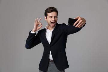 Image of joyous businessman 30s in formal suit showing ok sign while taking selfie photo on cell phone, isolated over gray background