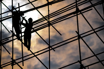 Silhouette of two construction workers wearing hard hats on scaffolding against the sun on a cloudy day 