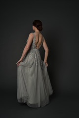 full length portrait of brunette girl wearing beautiful long gown, standing pose with background to the camera on grey studio background.