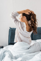 brunette woman making pony tail while sitting on bed in the morning