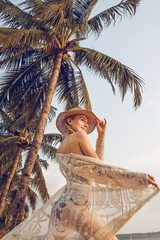 A beautiful young blond woman wearing a bikini and hat, around herself has a blanket, dancing on a sandy beach with a nice sky under the big palm trees at sunset.