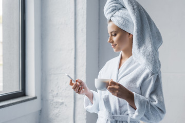 attractive young woman in bathrobe holding cup of coffee and using smartphone near the window - obrazy, fototapety, plakaty