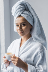 Obraz na płótnie Canvas Beautiful girl in bathrobe and white towel on head holding cup of coffee and looking at camera