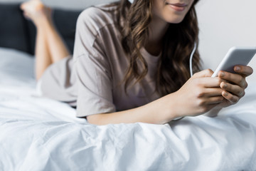 cropped view of woman listening music with headphones and smartphone while lying on bed