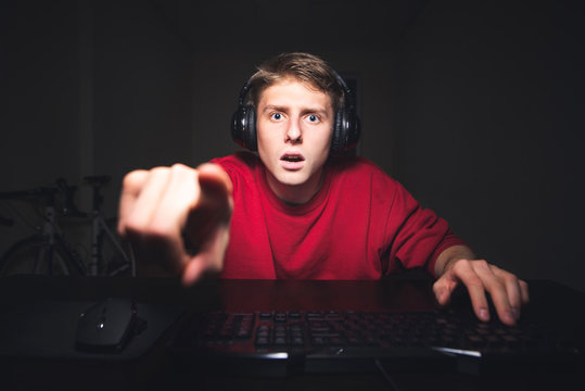 Surprised young man sits in the headphones at home in the room, uses a computer, looks into the camera and shows his fingers in the screen.