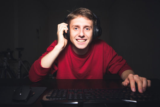 Portrait of a positive gamer at home for a computer game. Happy young man with a headset sitting at home at night, playing online video games, smiling and looking at the computer screen.
