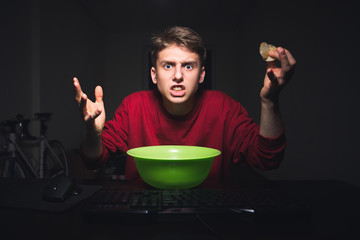 Young boy with the chips in his hand is looking at the computer screen with an outrage. Teenager is looking angrily at the monitor while watching videos on the Internet and eating snacks.