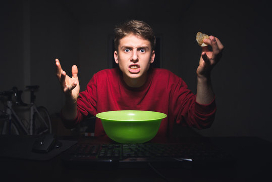 Young boy with the chips in his hand is looking at the computer screen with an outrage. Teenager is looking angrily at the monitor while watching videos on the Internet and eating snacks. - Powered by Adobe