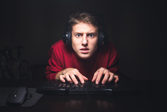 Focused gamer portrait looking at the game and using a computer keyboard. A concentrated young man in the headphones plays online home games on a computer and focuses on looking at the camera.