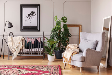 Grey armchair and plants in baby's bedroom interior with lamp next to cradle and poster. Real photo