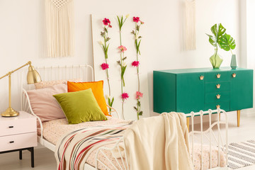 Olive green, orange and pastel pink pillow on bed with doted duvet and striped blanket, real photo of stylish bedroom interior with green cabinet and flower board
