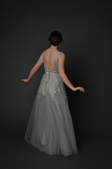 Fototapeta premium full length portrait of brunette girl wearing beautiful long gown, standing pose with background to the camera on grey studio background.