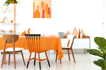 Three different chairs placed by the long dining table with orange tablecloth and dinnerware in...