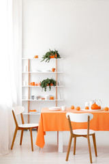 Real photo of white dining room interior with empty wall, rack with plants, cups and plates, wooden chairs and orange cloth on table with dinnerware