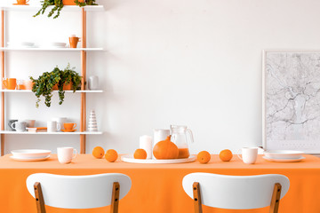 Real photo of two chairs placed by the table with mugs, bowls, plates, oranges, pumpkin and homemade juice in white dining room interior with map poster and rack with fresh plants