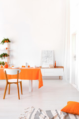 Long table with orange cloth, plates and fruits standing in white living room interior 