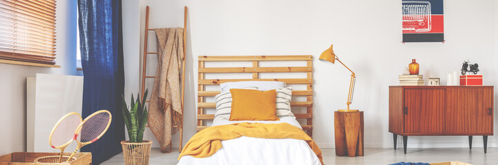 Bed with wooden bedhead, white sheets, ochre cushion and yellow knit blanket in real photo of white...