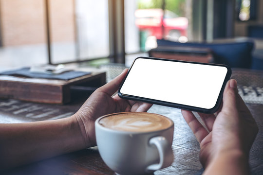 Mockup image of hands holding and using a black mobile phone with blank screen horizontally for watching with coffee cup on wooden table