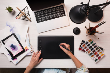 office desk with laptop, art supplies and cropped view of designer using graphics tablet and pen,...