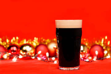 Dark ale beer in pint glass with christmas baubles tinsel and lights on red background