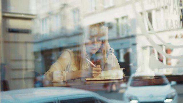 Young attractive blond woman looking outside through the window in a coffee shop smile write in notebook feel happy sitting cafe restaurant hand close up portrait slow motion