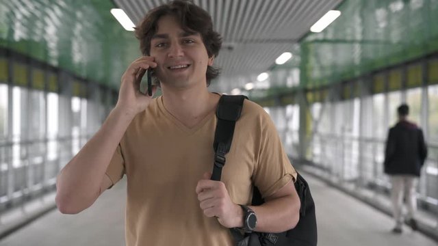 Attractive young caucasian man wearing beige t shirt and backpack is walking in tunnel, smiling and talking on smartphone. Tracking real time medium shot