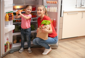 Young mother with daughter putting food into refrigerator at home after shopping